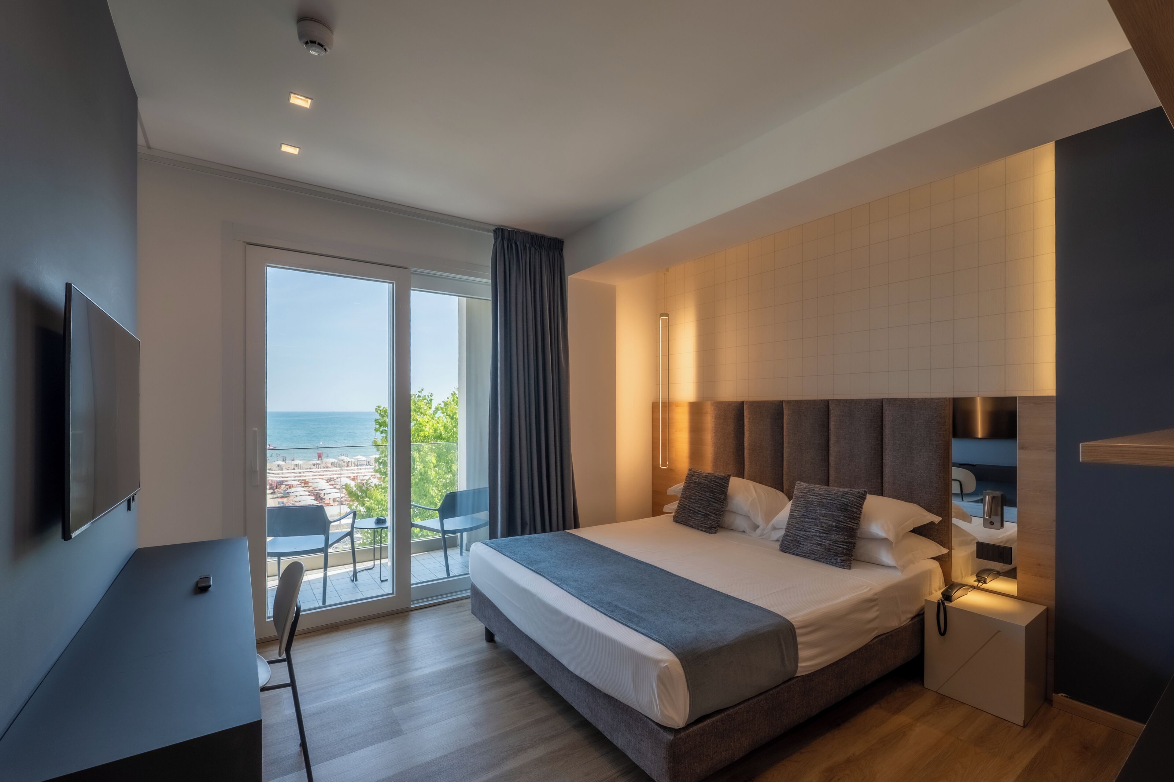 A modern hotel room with a large bed, a TV, a desk, and a view of the beach.