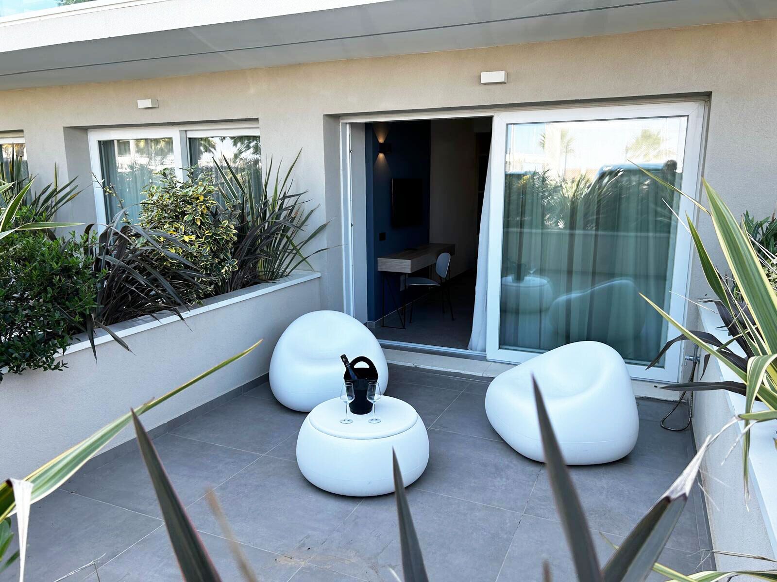 A modern outdoor seating area with white chairs and a table, surrounded by plants and a glass door leading to a room.