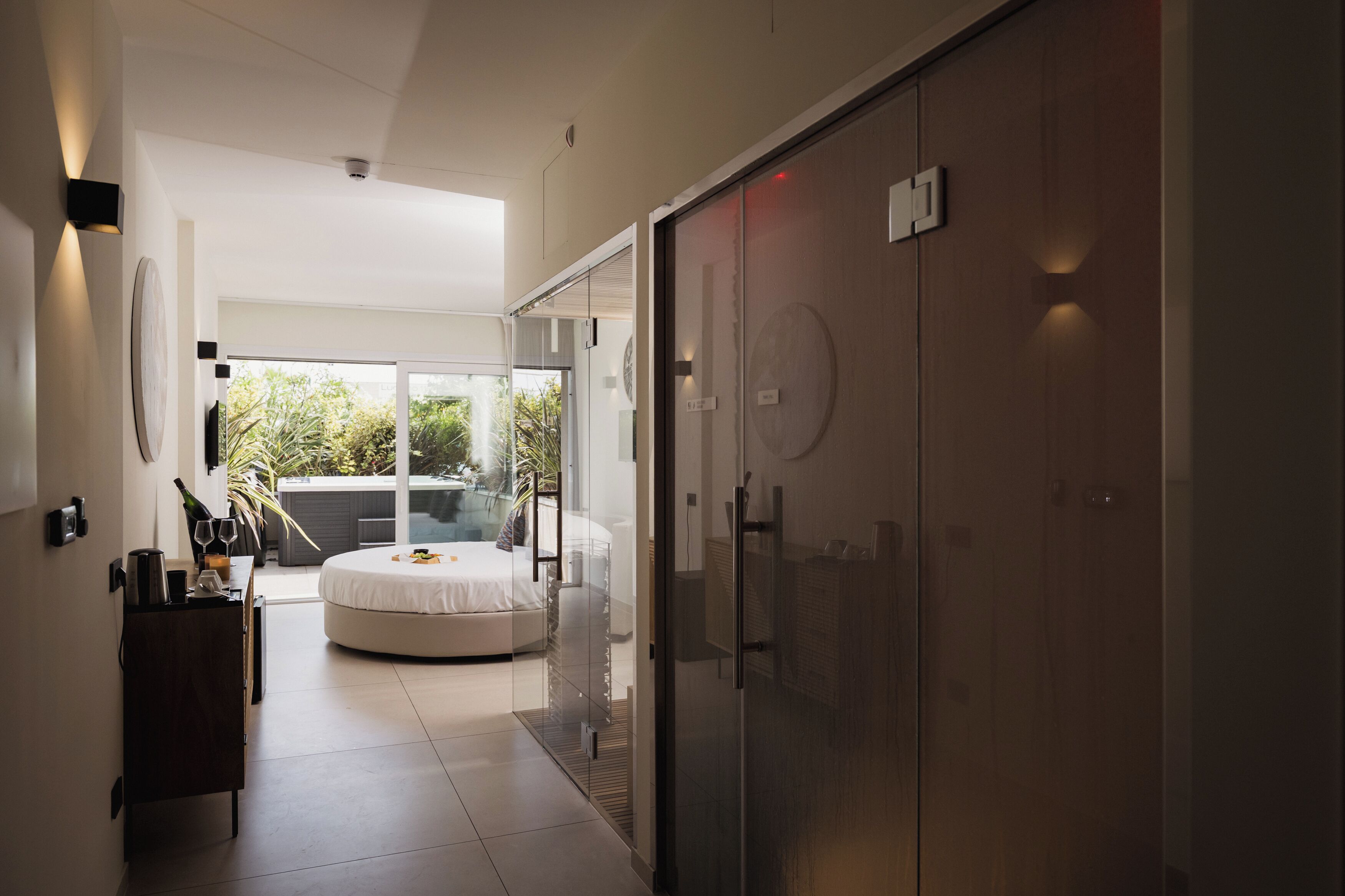 A modern hotel room with a bed, a glass door leading to a balcony, and a minibar.