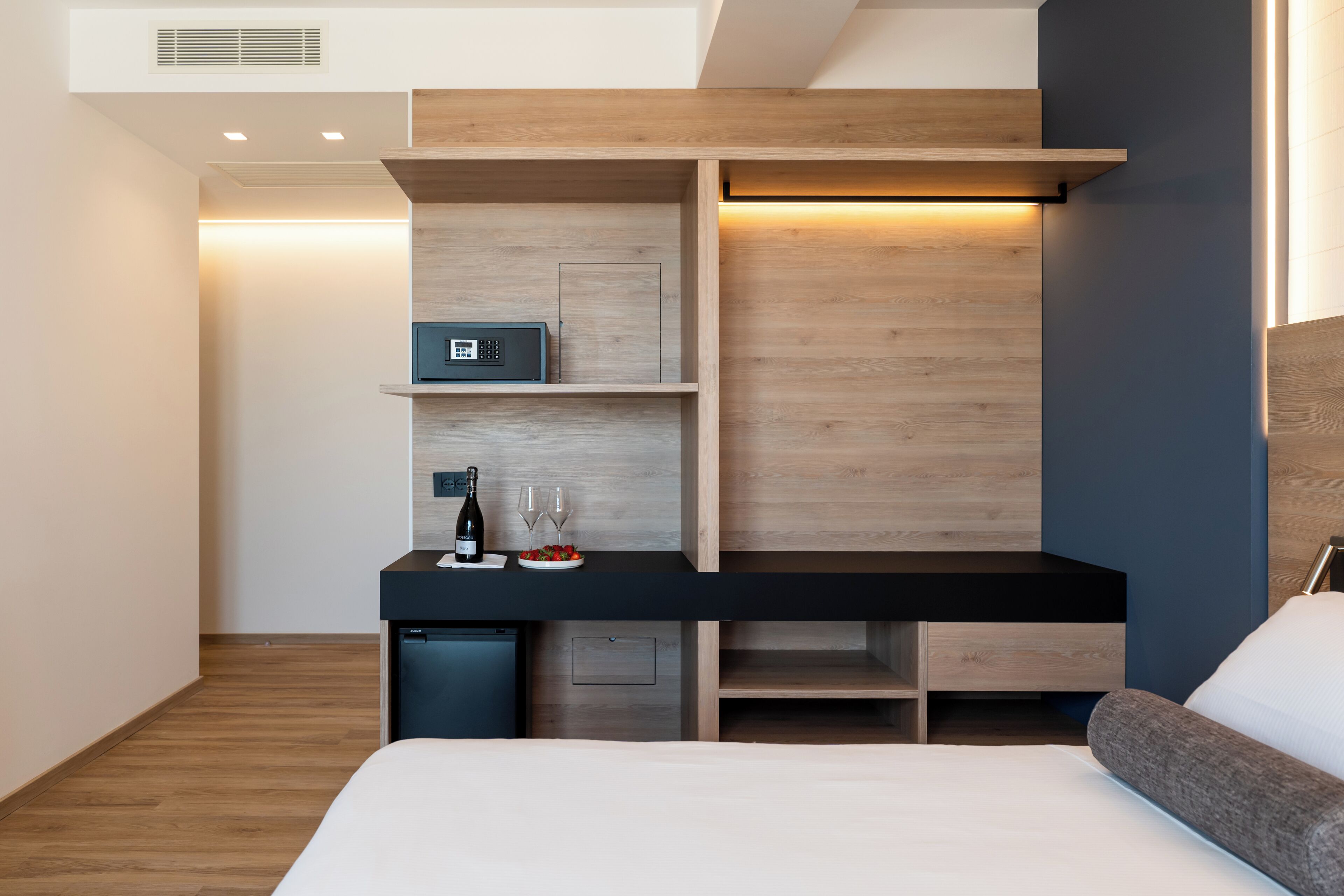 Una habitación de hotel moderna con un escritorio de madera, un refrigerador pequeño, una cama con cabecera gris y un estante en la pared con una botella de vino y copas.