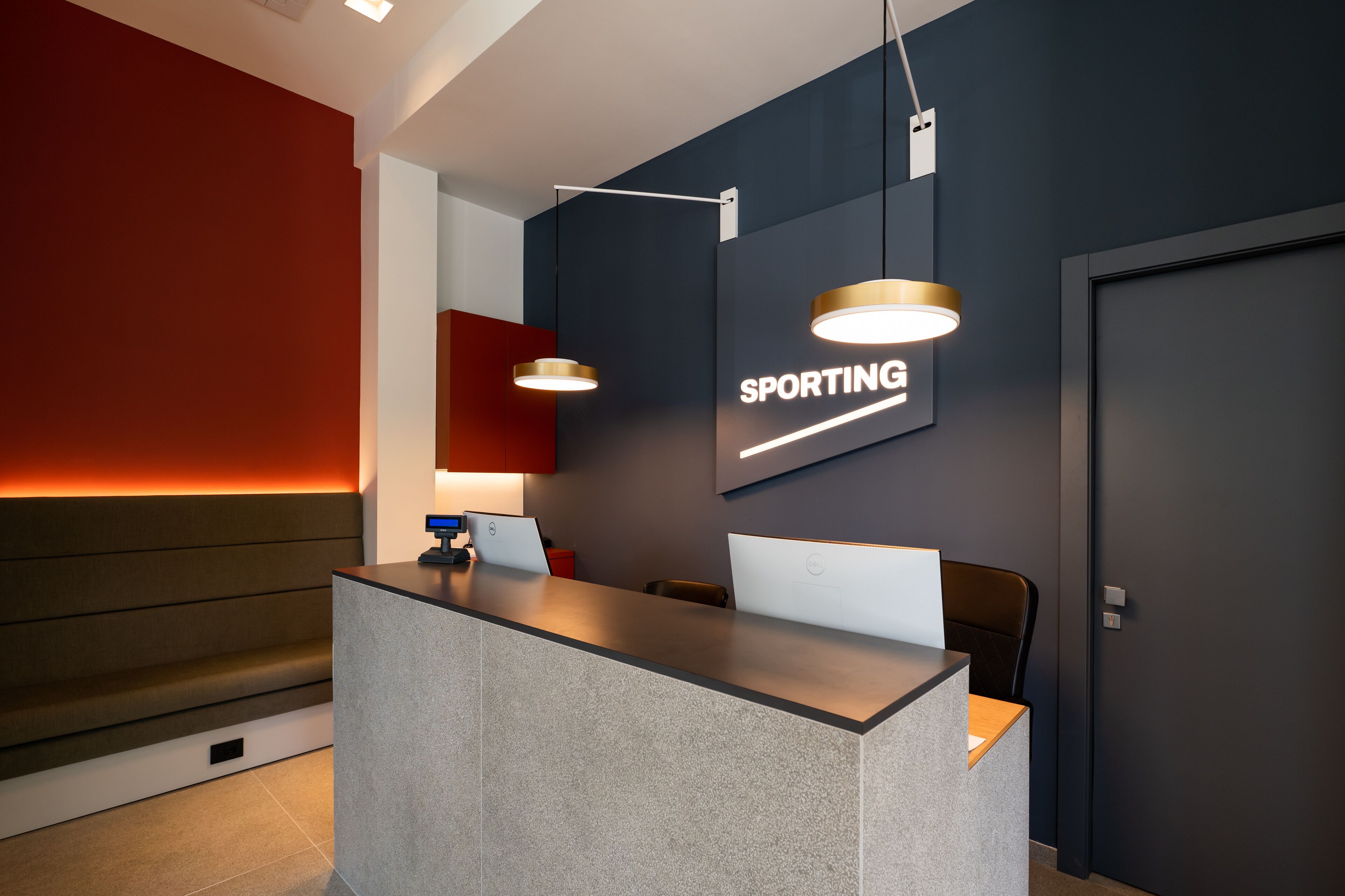 A modern reception desk with two laptops, a framed sign reading "SPORTING", and pendant lighting.
