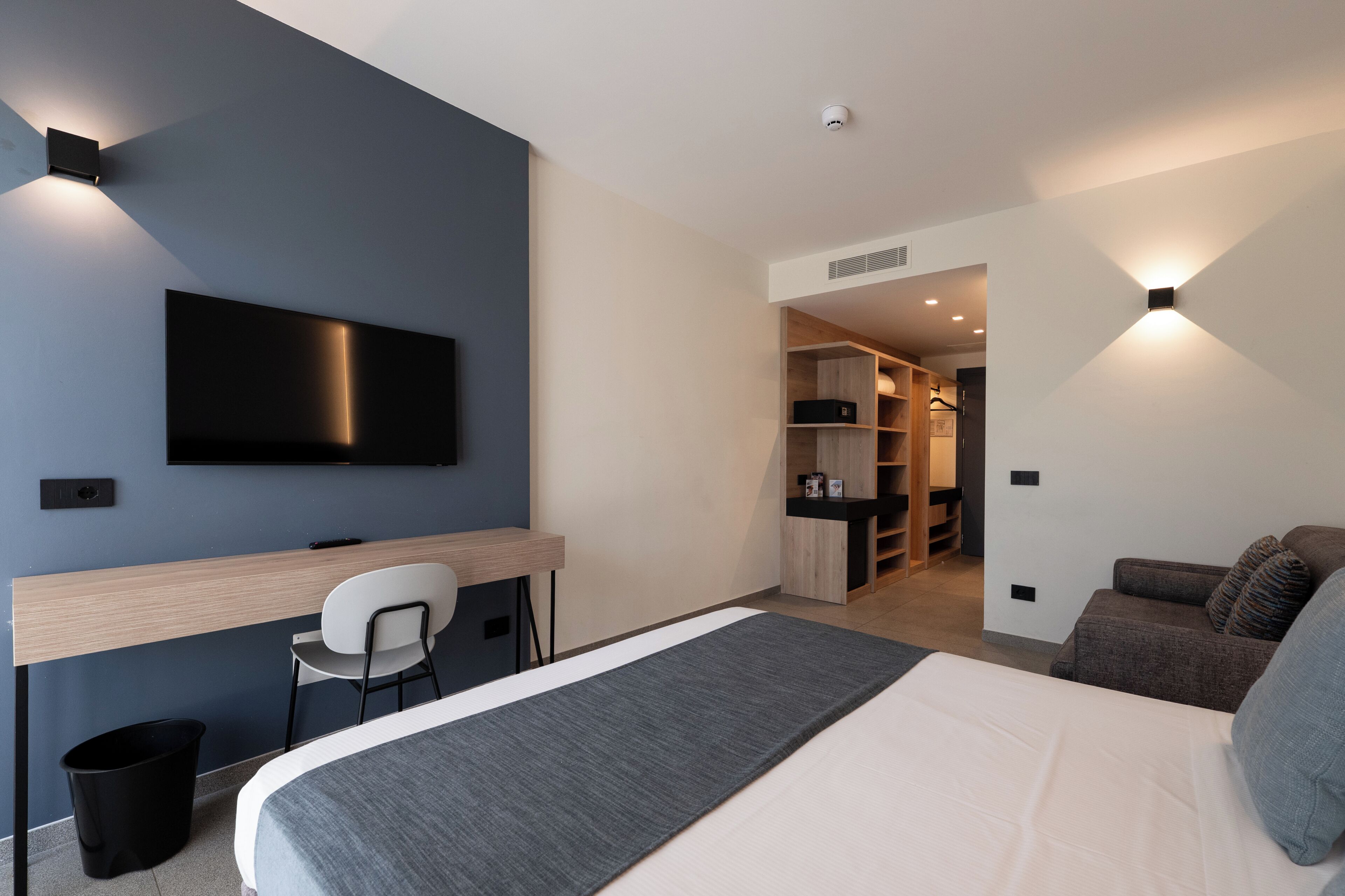 Moderni hotellihuone, jossa on sänky, työpöytä, tuoli, televisio ja pieni keittiö.