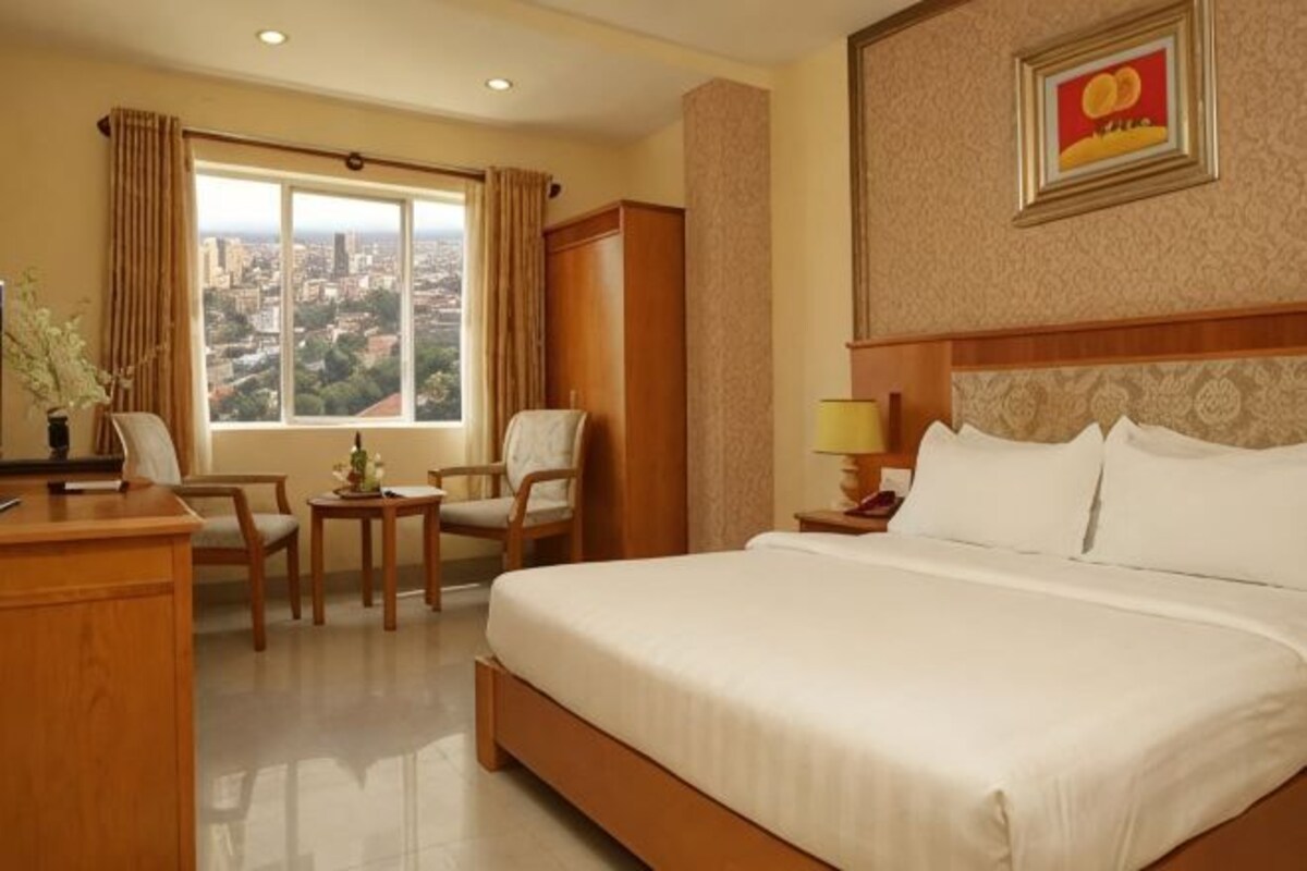 A hotel room with a large bed, a desk with a chair, a small table with a lamp, and a window offering a city view.