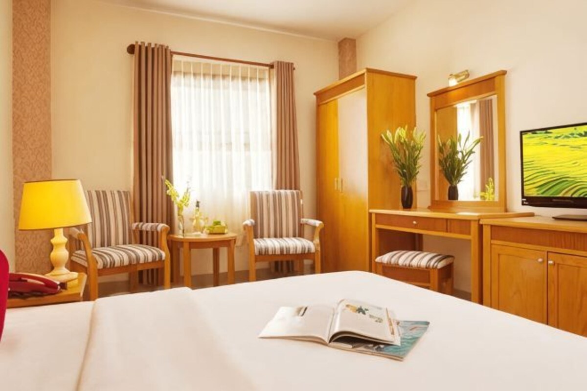 A hotel room with a bed, a TV, a desk with a chair, a small table with a lamp, and two chairs.
