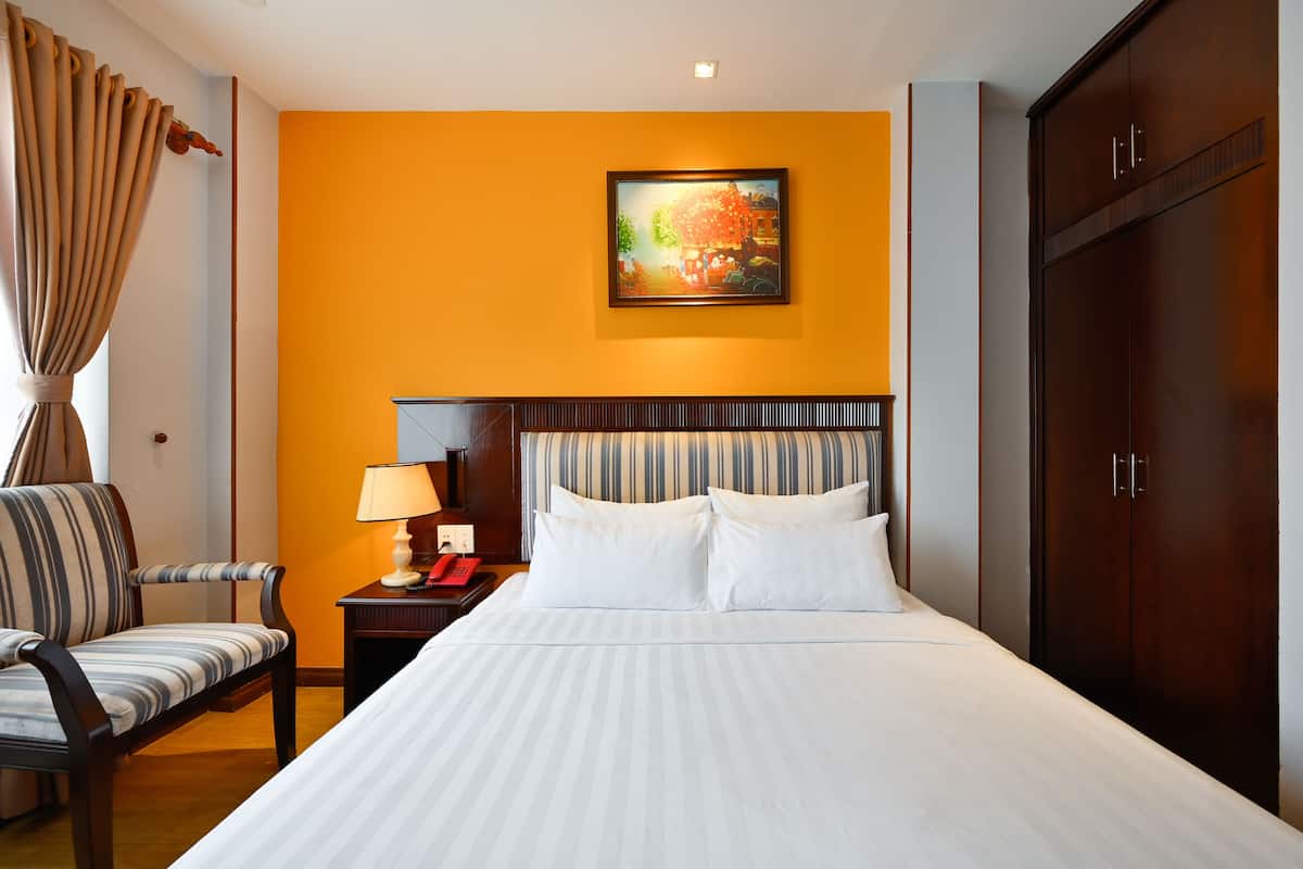 A hotel room with a bed, a chair, a nightstand with a lamp, a painting on the wall, and a wardrobe.