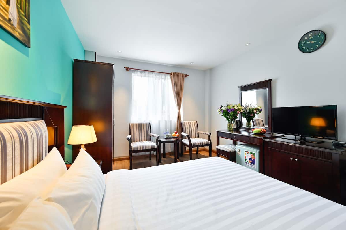 A hotel room with a bed, a desk with a TV, a chair, a lamp, and a window with curtains.