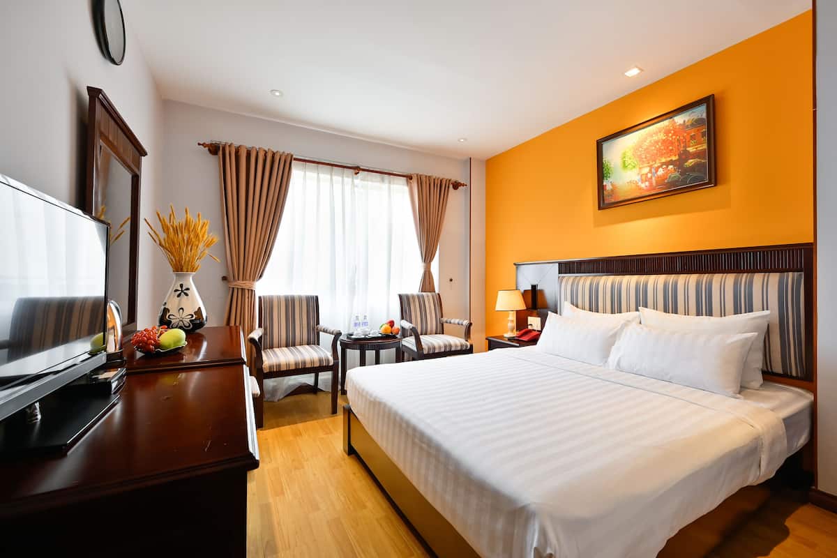 A hotel room with a large bed, a desk with a TV, two chairs, a small table with fruit, and a painting on the wall.