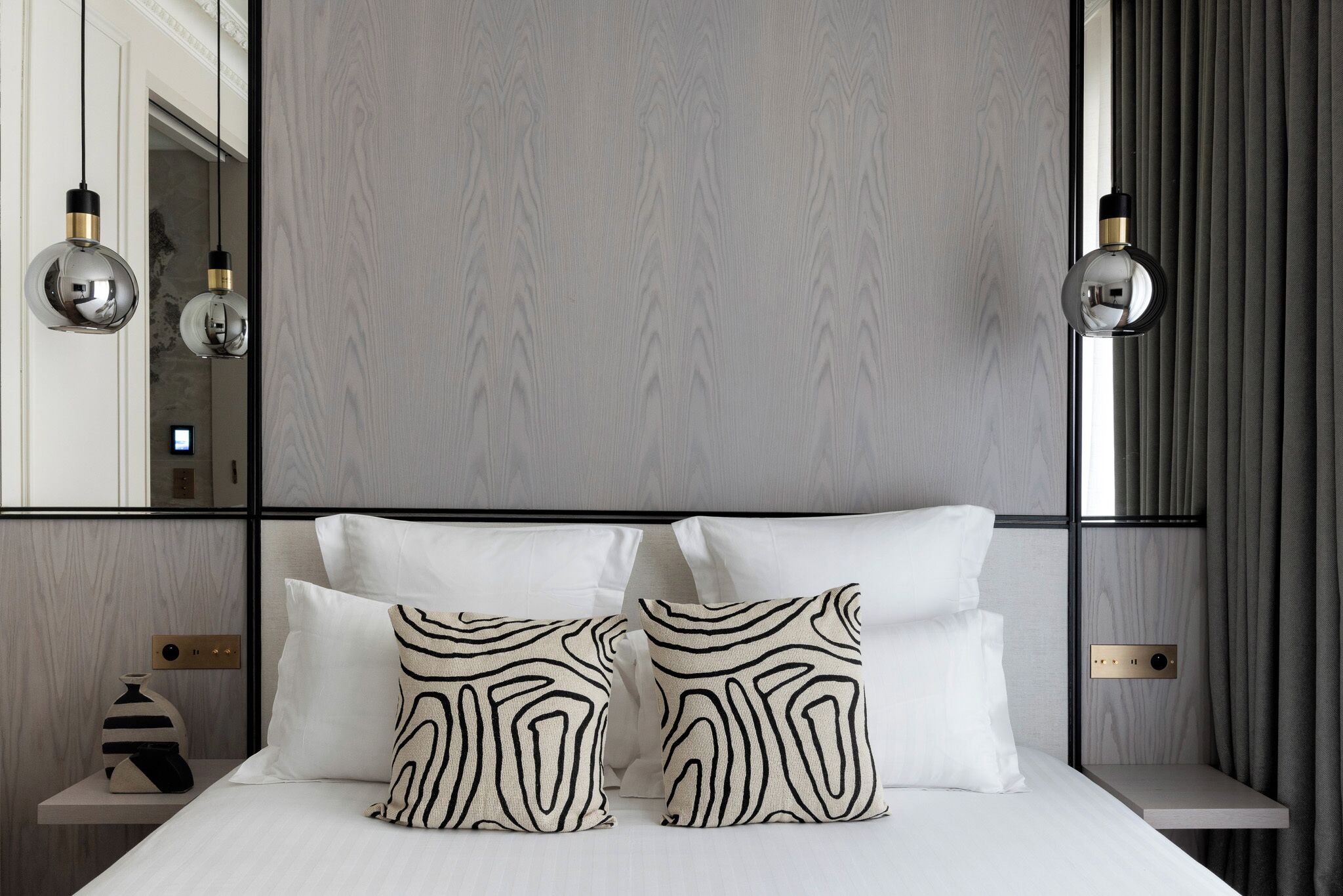 A modern hotel room with a large bed, two patterned pillows, a nightstand with a vase, and a mirror above the bed.