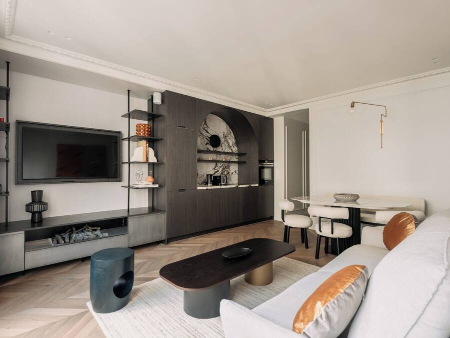 Una sala de estar moderna con un televisor de pantalla plana, una mesa de centro oscura, un sofá con cojines, un comedor con mesa y sillas, y una cocina con salpicadero de mármol.