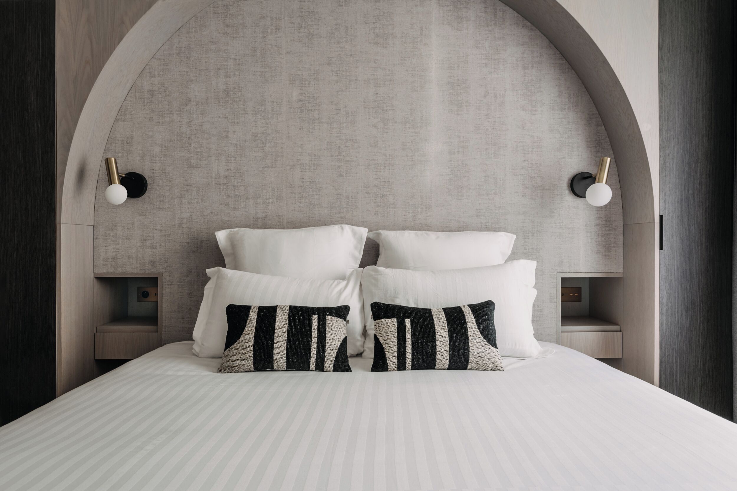 A bed with white bedding and black and white striped pillows, flanked by two bedside tables with lamps, set against a textured wall with an arched design.