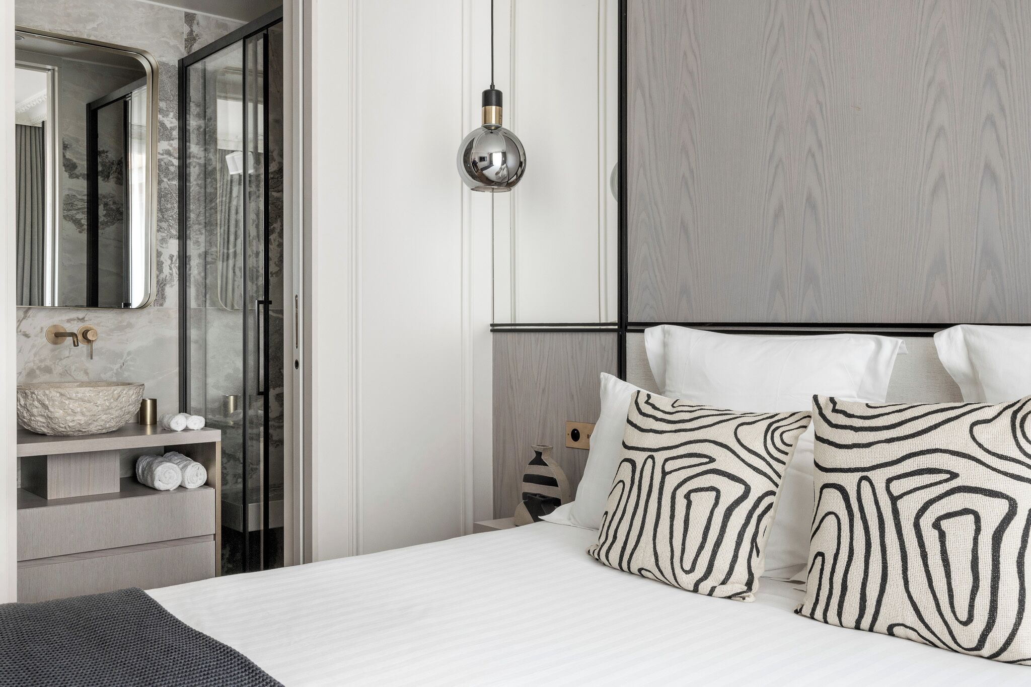 A modern bedroom with a large bed, a bedside table with a mirror, and a wall-mounted light fixture.
