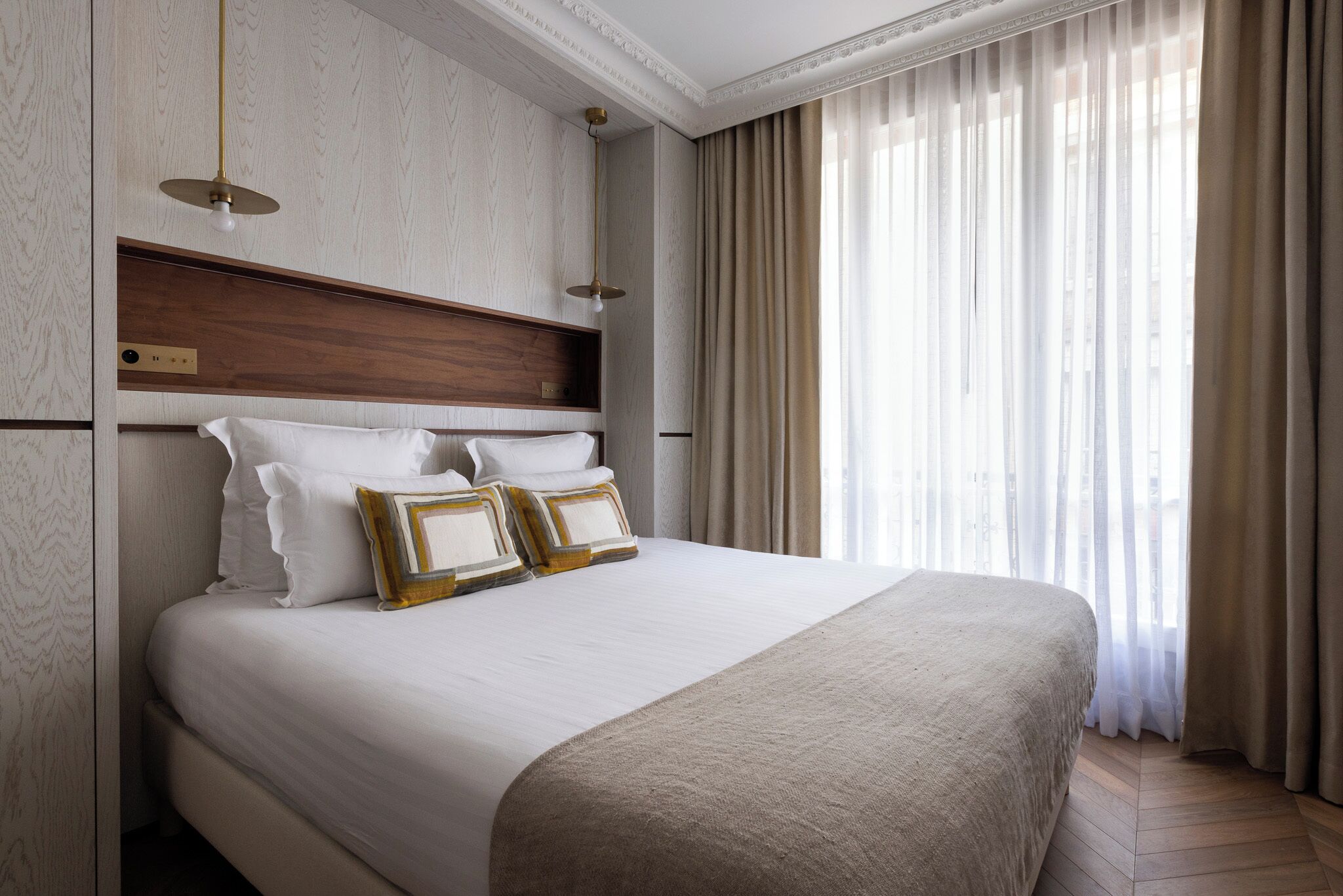 A large bed with white linens and a beige throw blanket, a wooden headboard, and a pendant light.