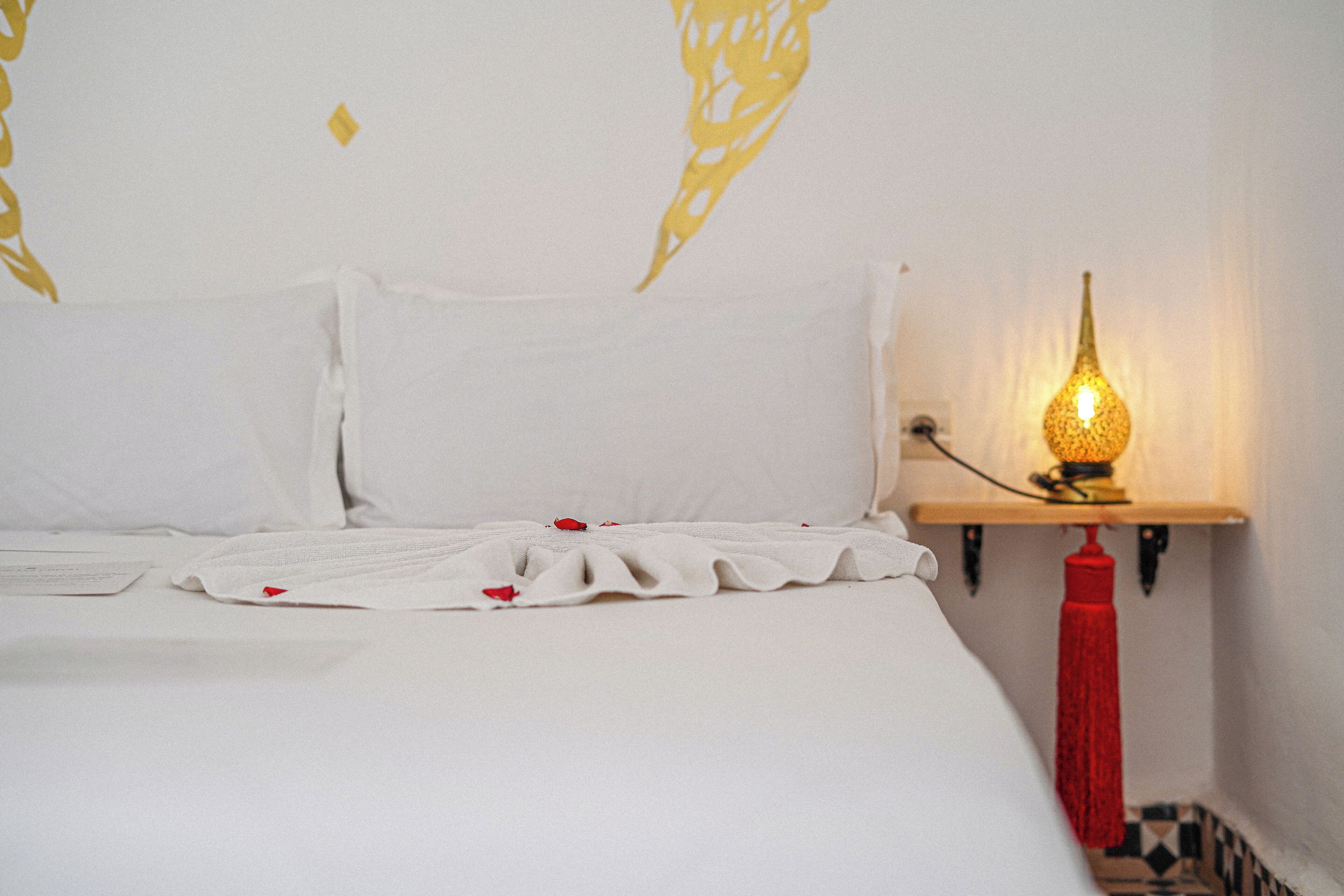 A neatly made bed with white linens and a red tassel lamp on a bedside table.