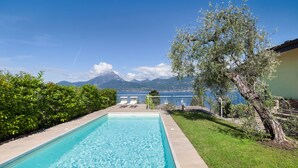 Ein rechteckiger Swimmingpool mit klarem, blauem Wasser, umgeben von einer gepflegten Rasenfläche und einem Baum. Im Hintergrund sind ein Berg und ein Gewässer zu sehen.