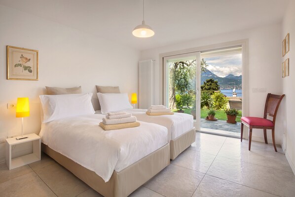 A bedroom with two beds, a chair, and a view of the outdoors. - Villa Palladini With Pool (Torri del Benaco)