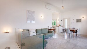 Ein modernes Wohnzimmer mit Glastrennwand, einer Couch, einem Esstisch mit Stühlen und einer Küchenzeile im Hintergrund.