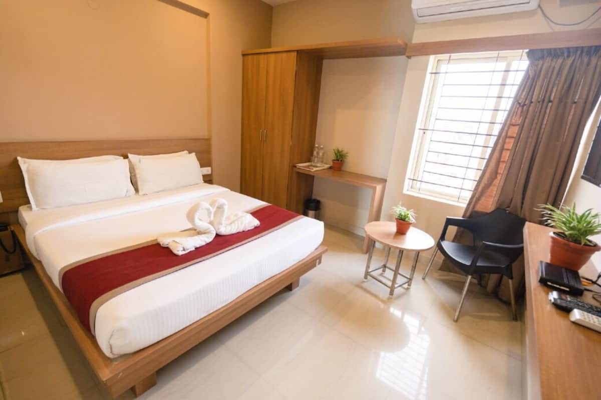 A hotel room with a bed, a desk with a chair, a small table with a chair, a wardrobe, and a window with curtains.