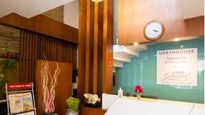 A reception area with a wooden reception desk, a clock, and a sign for Urbanhouse Samash Rooms & Suites by Live Grans Hotels. - Samasth Rooms and Suites by YGH (Mysore)