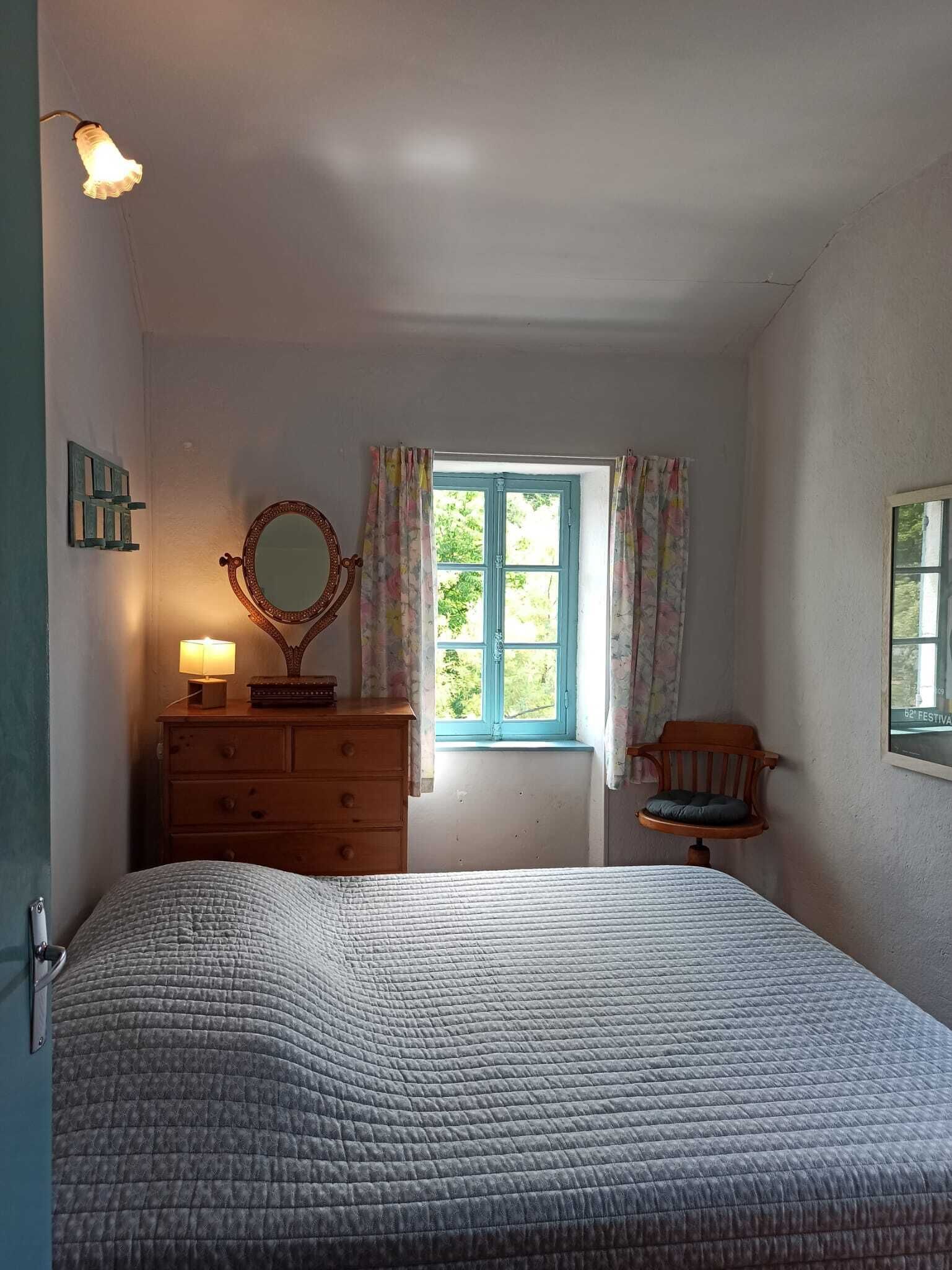 Een slaapkamer met een bed, een dressoir, een spiegel, een raam met gordijnen en een stoel.