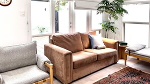 A living room with a beige sofa, a grey armchair, and a patterned rug. There are two windows with blinds, a potted plant, and a clock on the wall. - Sandstone Villa In City Fringe Location (Fullarton)