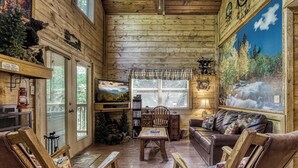A cozy cabin interior with wooden walls, a fireplace, a dining table, and a large window with a view of trees.