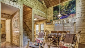 A log cabin interior with a staircase, rocking chairs, a dining table, and a large window with blinds.