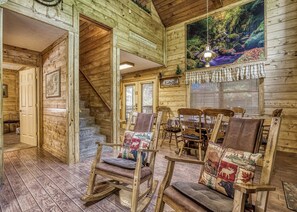 L’intérieur d’une cabane en rondins, avec un escalier, des fauteuils à bascule, une table à manger et une grande fenêtre équipée de stores.