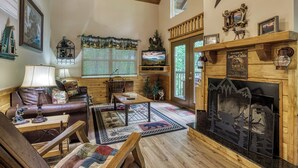Interior acogedor de una cabaña con chimenea, muebles de madera, televisor y vista panorámica por la ventana.