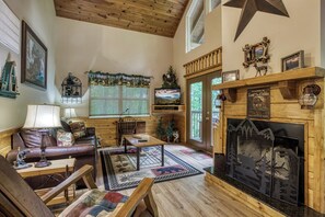 Interior acolhedor de uma cabana com lareira, móveis de madeira, televisão e vista panorâmica pela janela.