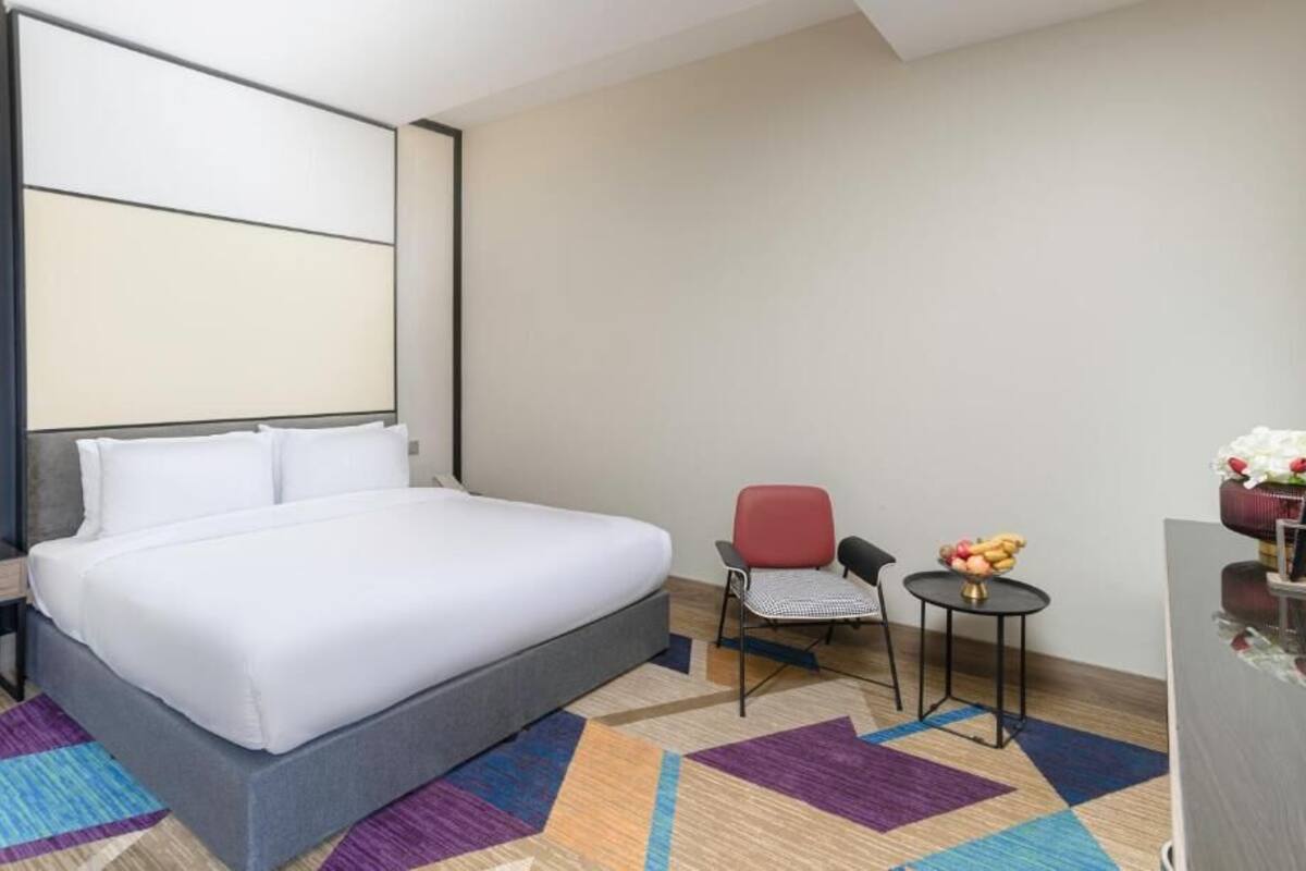 A hotel room with a large bed, a small table with a fruit bowl, a chair, and a decorative vase with flowers.