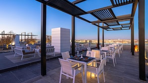 Eine Terrass mit weißen Stühlen und Tischen, einer Pergola und einem Blick auf die Stadt im Hintergrund.