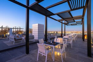 An outdoor terrace with white chairs and tables, a pergola structure, and a city view in the background.
