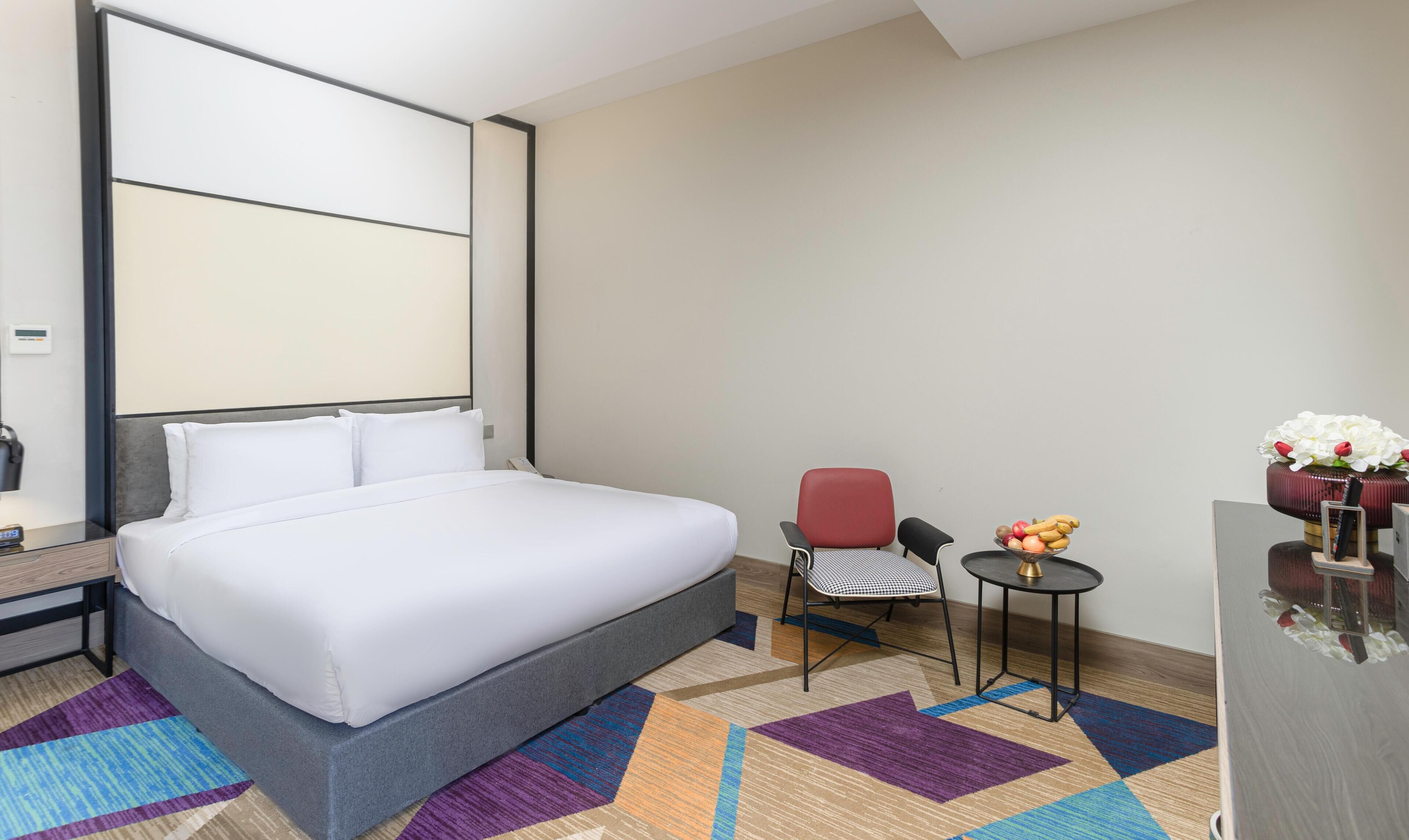 A modern hotel room with a large bed, a desk, a chair, a small table with fruit and flowers, and a decorative vase.