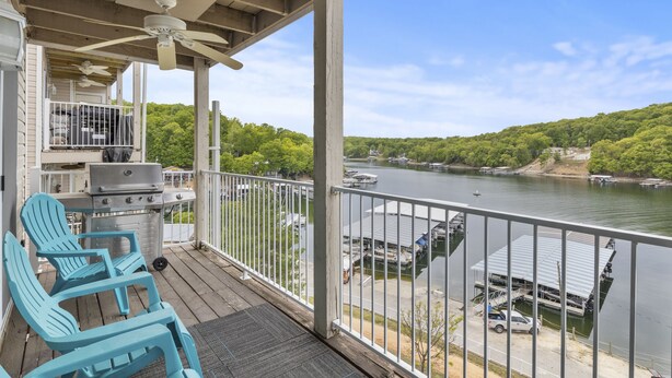 A covered deck with a grill and chairs, overlooking a lake with boats and docked vehicles.
