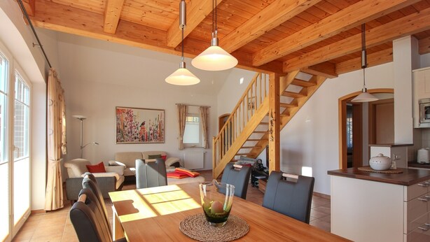 Een ruime woonkamer met houten plafond, een eettafel met stoelen, een bank en een trap naar een bovenverdieping.