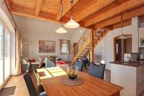 Amplia sala de estar con techo de madera, una mesa de comedor con sillas, un sofá y una escalera que sube a un nivel superior.