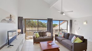 Relax in this spacious living area with a high ceiling, ceiling fan, and sliding glass door leading to a balcony with a view of palm trees and a white fence.