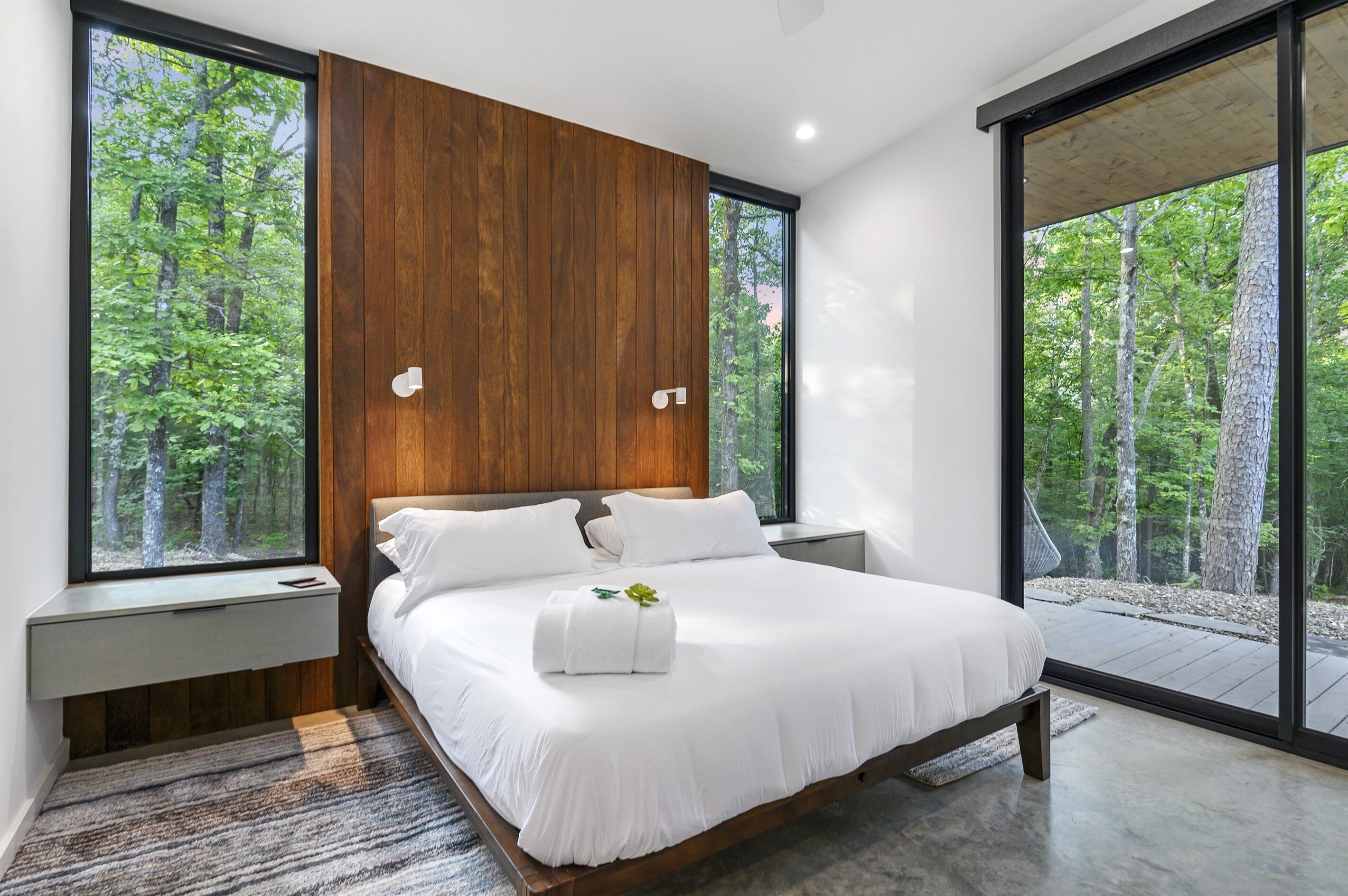 Een moderne slaapkamer met een groot bed, een wand van houten panelen en grote ramen met uitzicht op een bos.