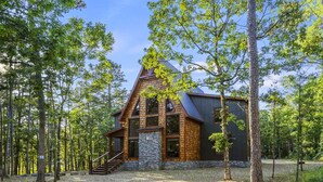A cabin with a stone foundation, wooden exterior, and a steeply pitched roof, surrounded by tall trees. - Family Retreat with Game Room and Playset (Broken Bow)