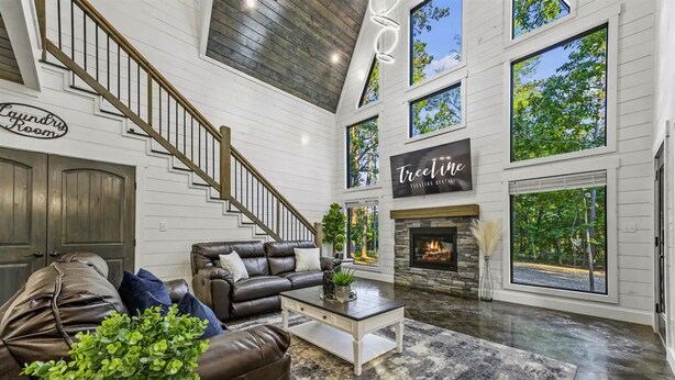 A spacious living room with a leather sofa, a coffee table, and a fireplace. There is a staircase with a sign reading "Laundry Room" and a sign for "TreeLine Fitness Retreats." - Family Retreat with Game Room and Playset (Broken Bow)
