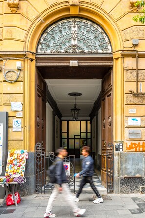 L’entrée d’un immeuble, ornée d’un portail en fer forgé, une pancarte indiquant « À LOUER », et un vendeur ambulant présentant une variété d’articles colorés.