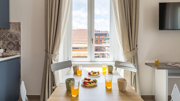 A dining area with a table set for two, a TV mounted on the wall, and a window with curtains.