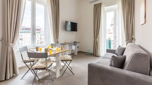 A modern living room with a sofa, a dining table with chairs, a flat-screen TV mounted on the wall, and large windows with curtains.