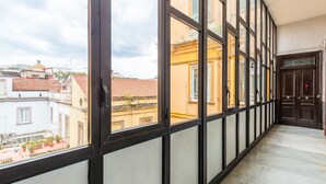 A corridor with large windows, a tiled floor, and a view of buildings with a dome and red-tiled roofs.