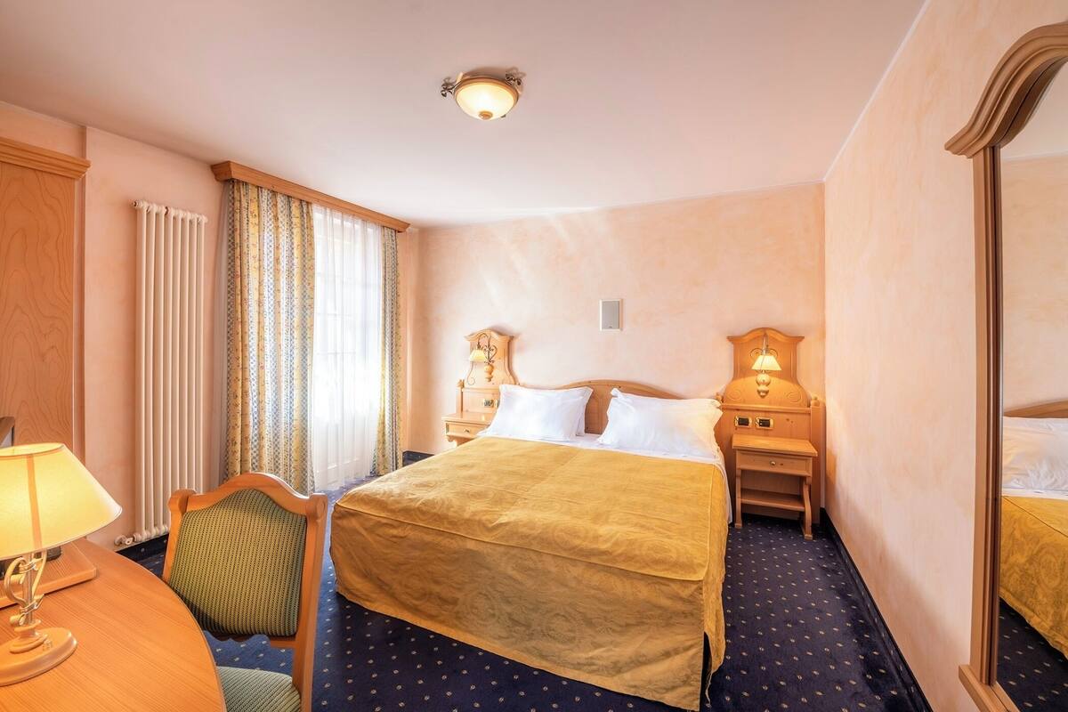 Ein Hotelzimmer mit einem großen Bett, einem Sessel, einem Schreibtisch mit Lampe, einem Nachttisch ebenfalls mit Lampe und einem Spiegel.