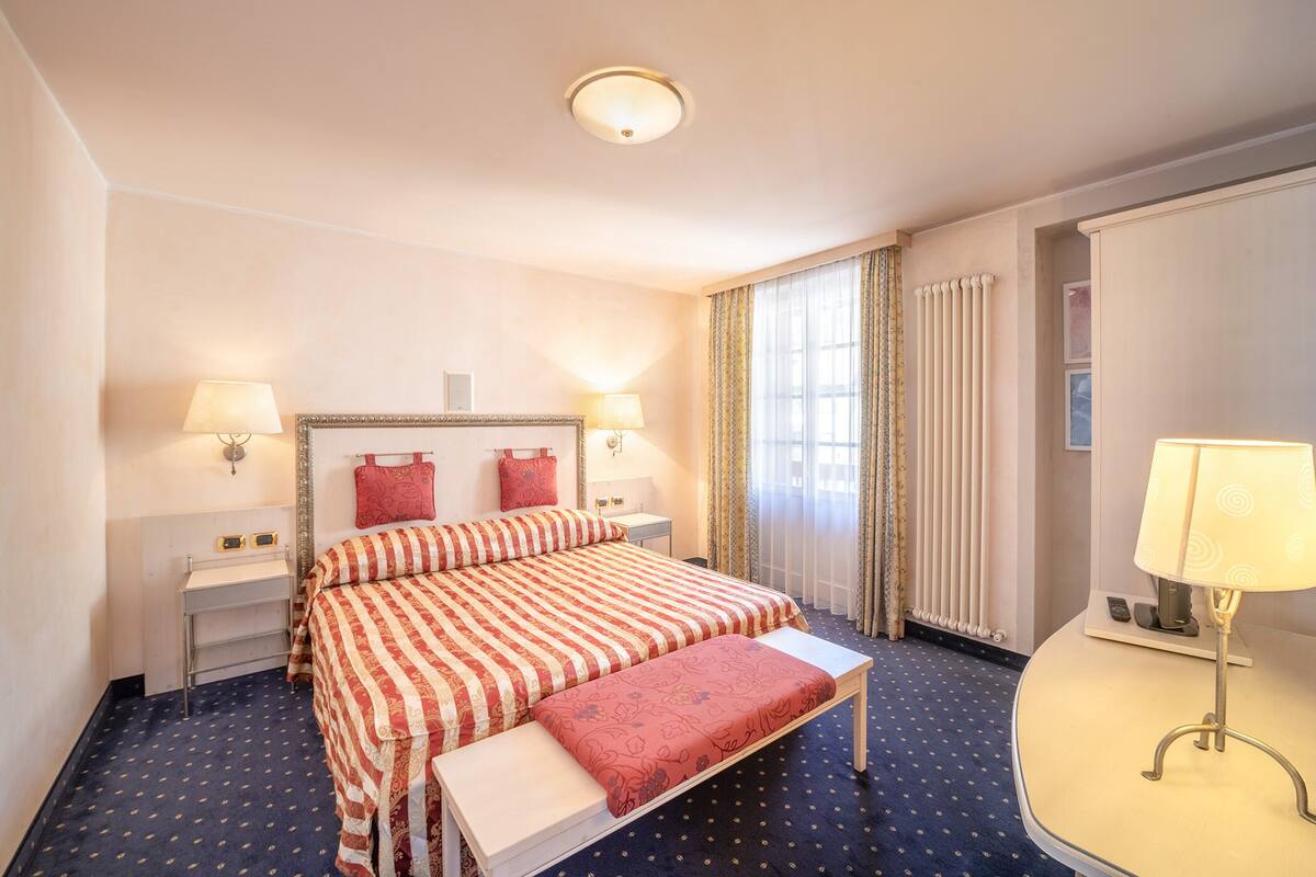 Ein Hotelzimmer mit einem Bett, Nachttischen, einer Bank, einer Lampe und einem Fenster mit Vorhängen.