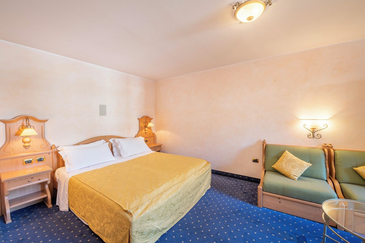 Une chambre d’hôtel avec un lit, une table de chevet, un canapé vert et des lampes fixées au mur.