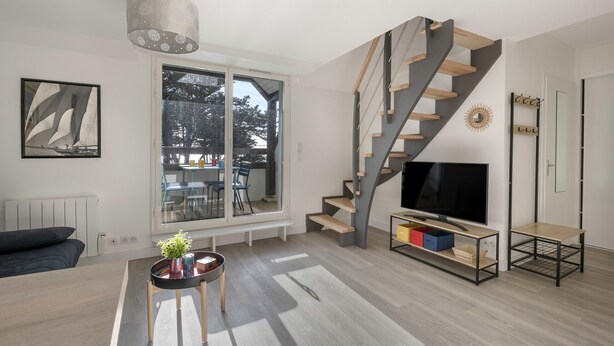 Ein modernes Wohnzimmer mit großem Fenster, einer Treppe, einem Fernseher und einem kleinen Tisch mit einer Pflanze.