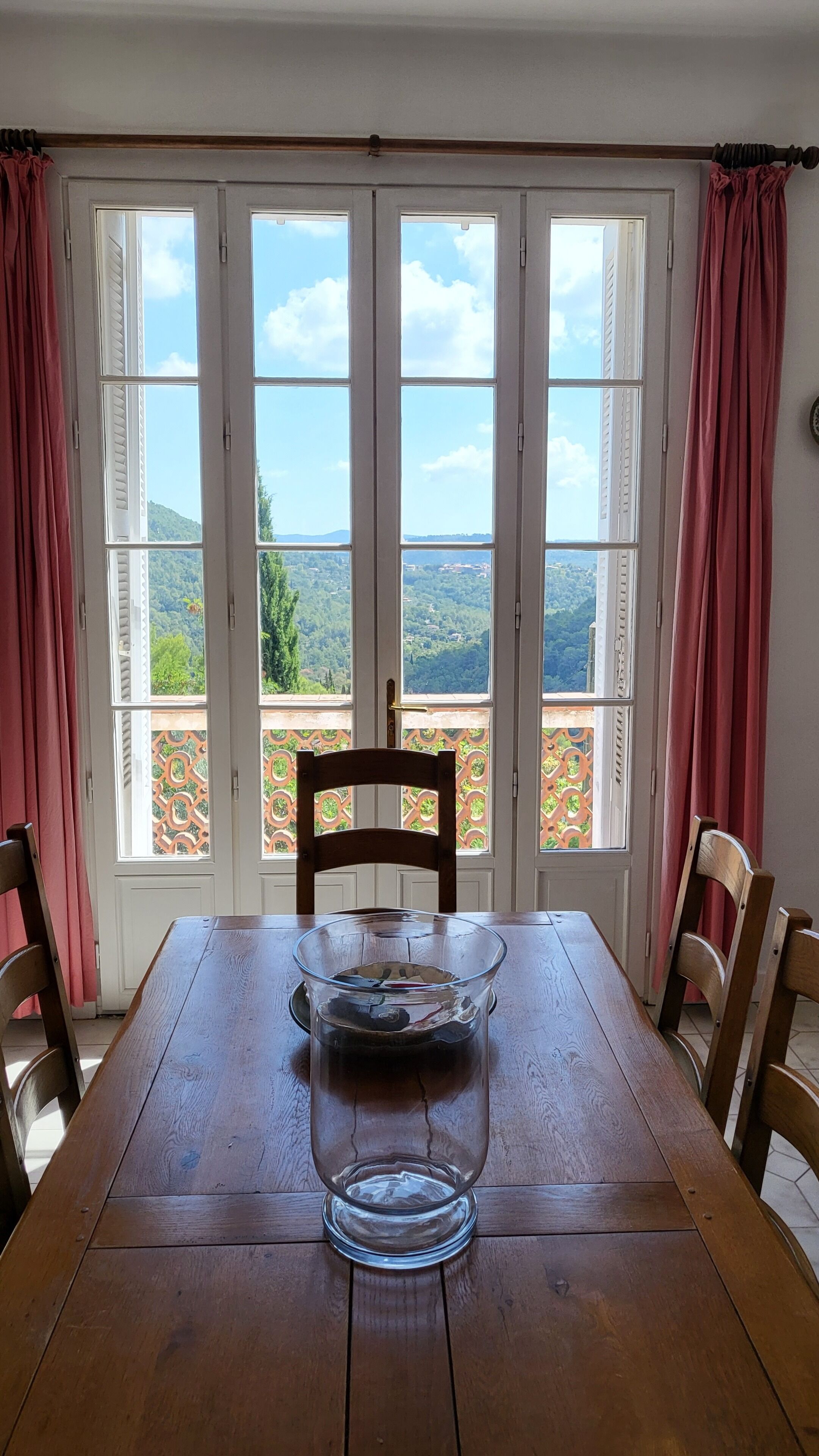 A dining table with a glass vase in the center, set against a backdrop of large windows with red curtains, offering a view of a lush landscape.