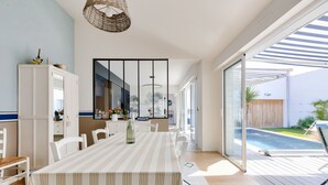 Ein geräumiger Essbereich mit einem langen Tisch, weißen Stühlen und einer Tischdecke mit Streifen. Über dem Tisch hängt eine Lampenschirm aus Weidengeflecht und an der Wand ein großes Spiegelbild.